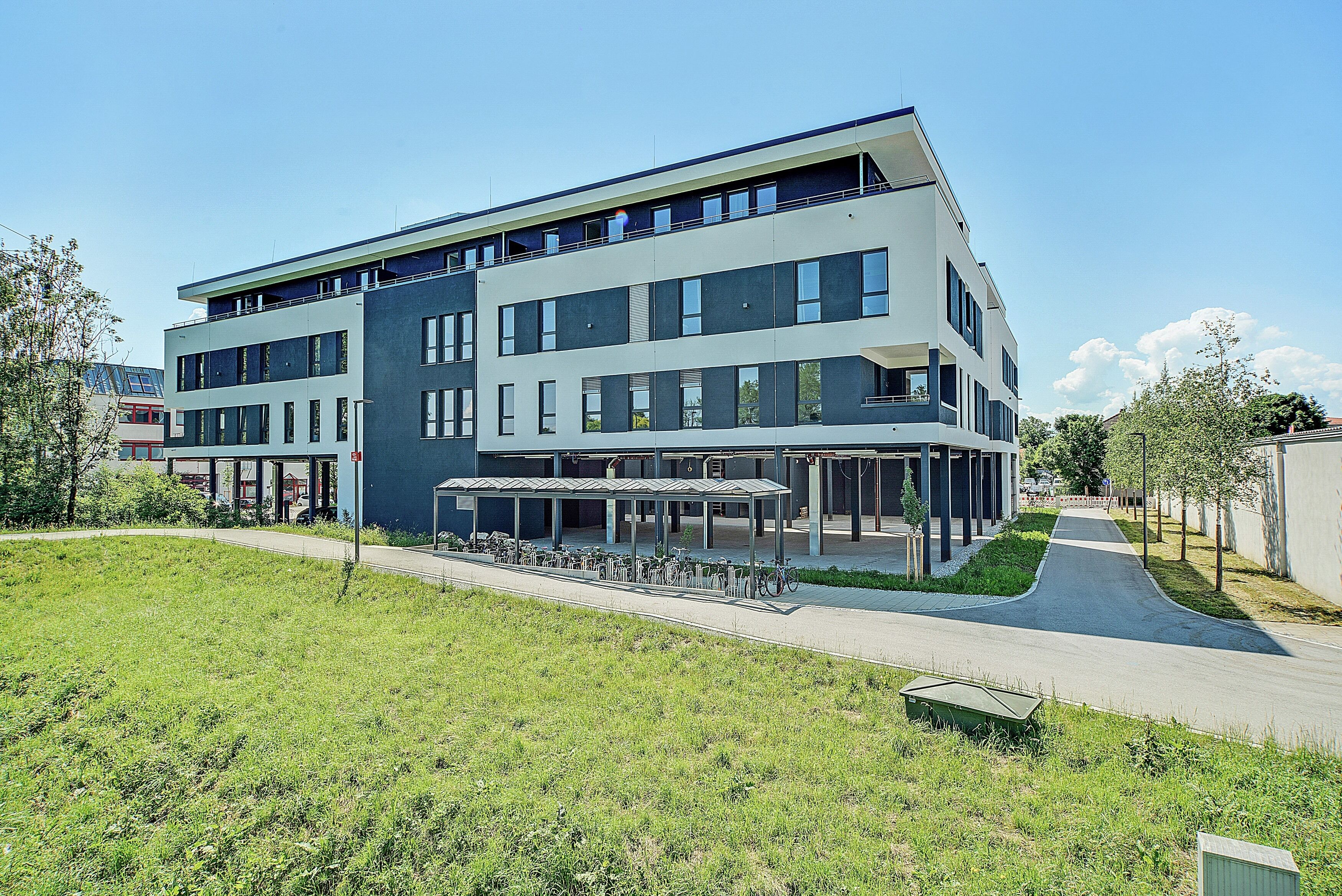 A modern multi-story building with a covered outdoor seating area and a pathway leading to the entrance.