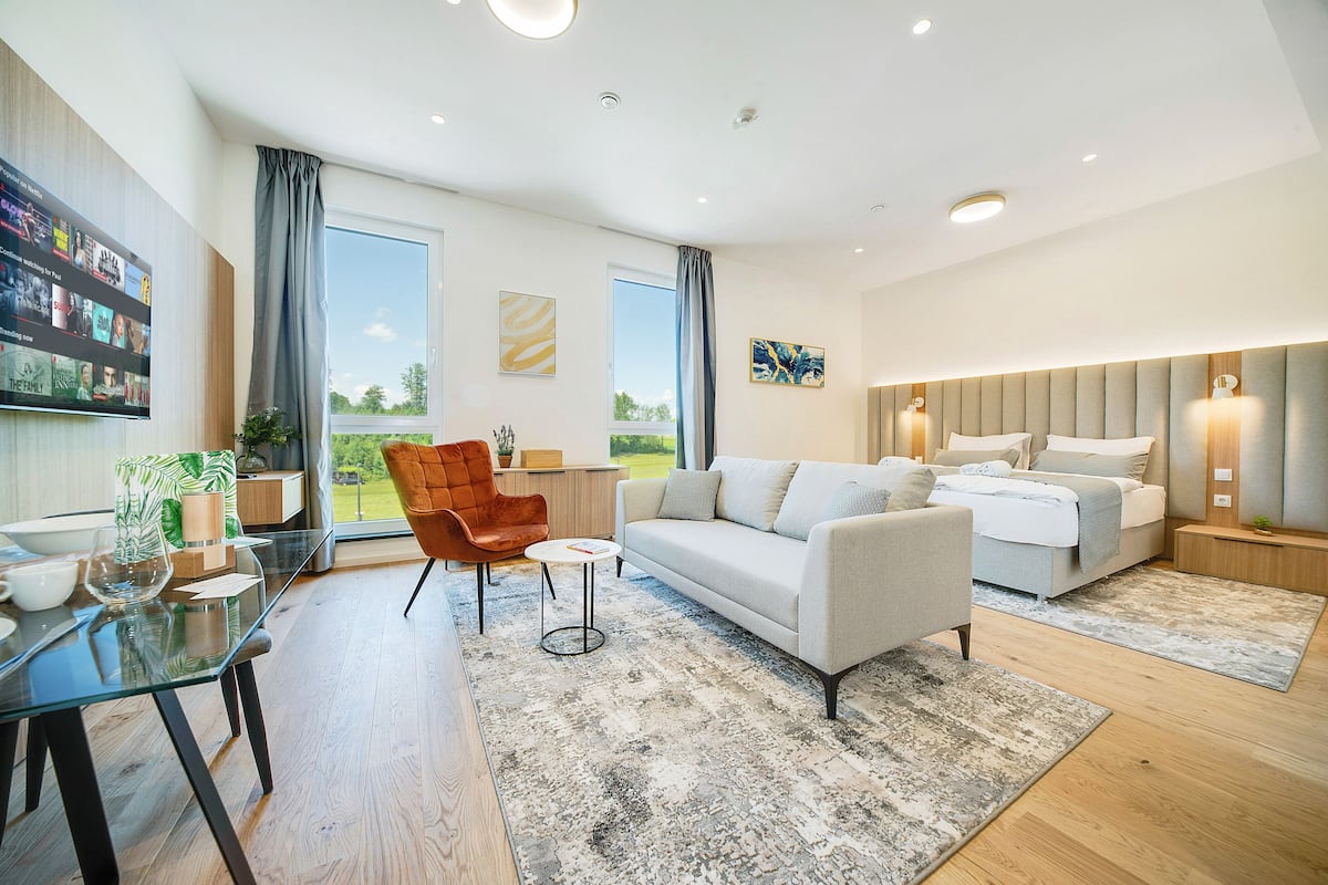 A modern living room with a sofa, armchair, and coffee table, a dining area with a glass table, and a large window with curtains.
