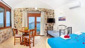 Une pièce avec un mur en pierre, un lit recouvert d’une couverture bleue, une table à manger avec des chaises, un téléviseur et une vue sur la mer.