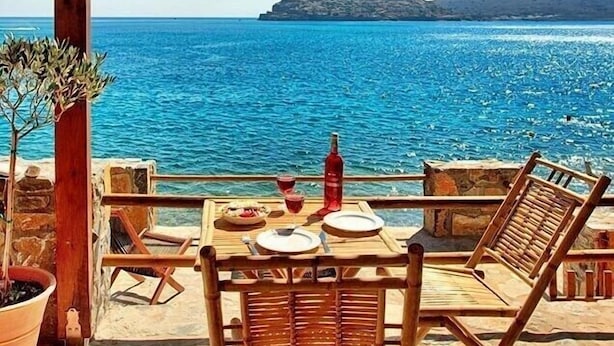Une pergola en bois, avec une table et des chaises, donnant sur une mer d’un bleu profond et sur une île lointaine.