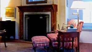 A cozy room with a fireplace, a bench, a table with a lamp, and a window with blinds. - Historic Cottage on Ware River Estate (Gloucester)