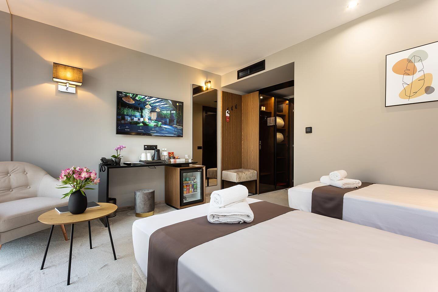 Une chambre d’hôtel avec deux lits, un canapé, une petite table ornée d’un vase de fleurs, une télévision, un minibar et une porte donnant sur une salle de bain.