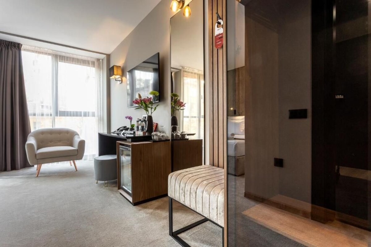 Una habitación de hotel moderna con sofá, escritorio con espejo, pequeña mesa con flores y un ventanal con cortinas.