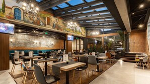 Een modern restaurant met een hoge plafond, zichtbare balken en een muurschildering. Er staan tafels en stoelen klaar voor het diner, en een televisie is aan de wand bevestigd.