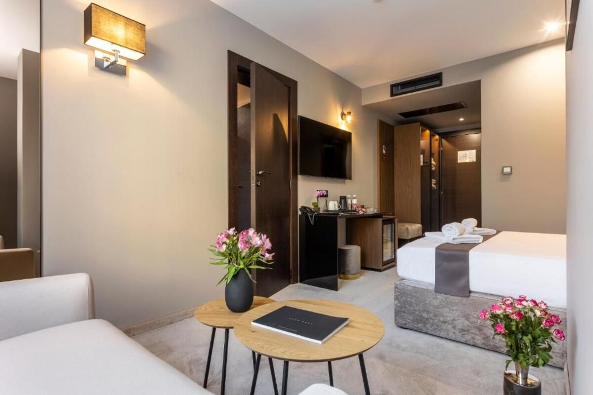 Una habitación de hotel con una cama, un escritorio, una silla, una mesita con un libro y un jarrón de flores.