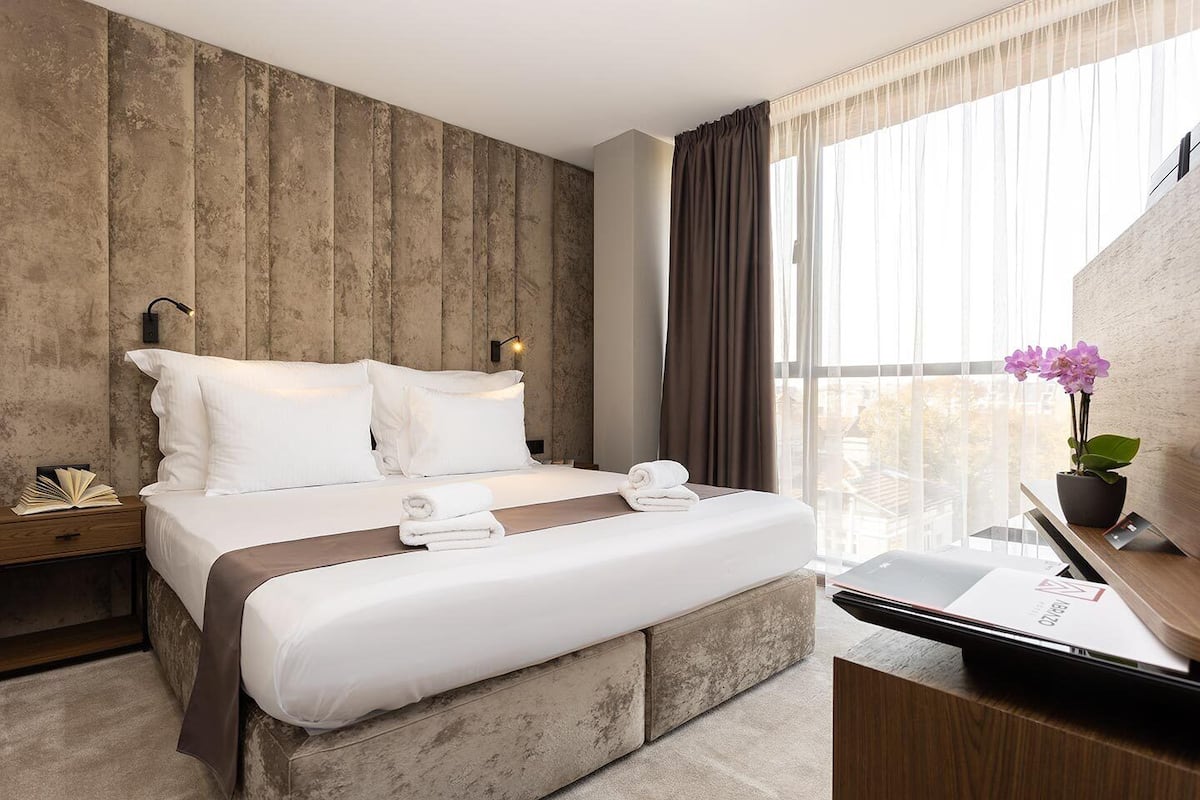 A modern hotel room with a large bed, a nightstand, a desk with a computer, and a view of the city through sheer curtains.