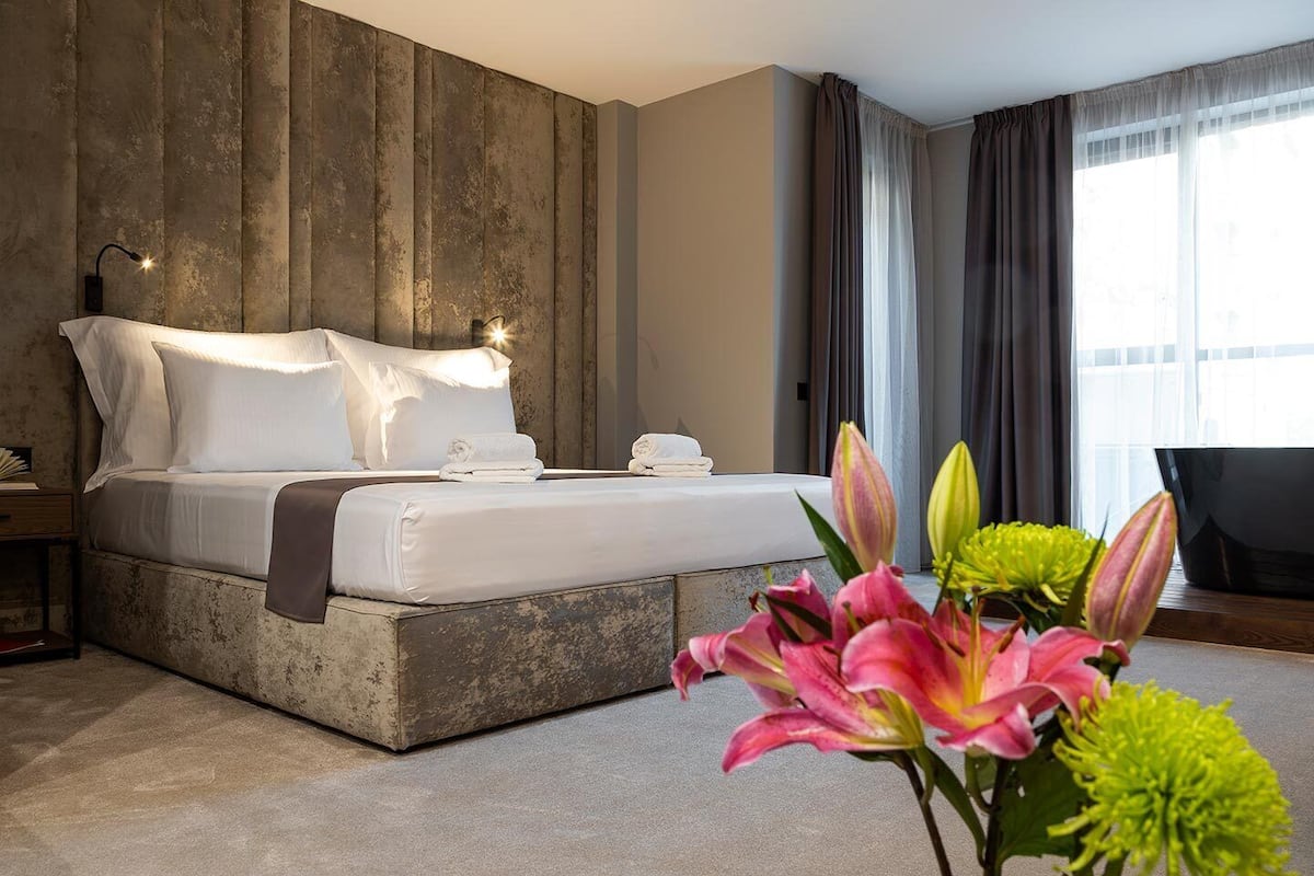 A modern hotel room with a large bed, a wooden accent wall, a desk with a lamp, and a view of the city through large windows.