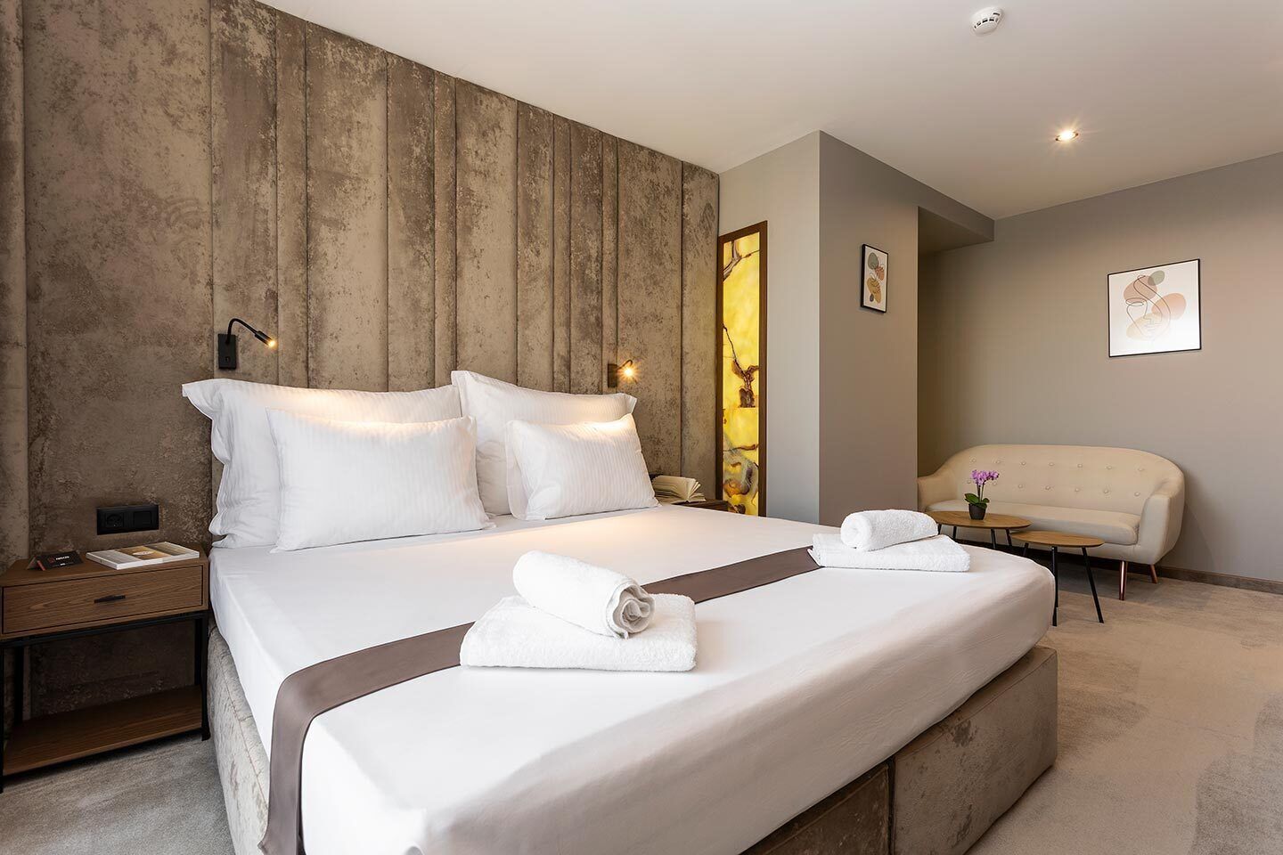A modern hotel room with a large bed, a bedside table with a lamp, a chair, and a small table with a plant.