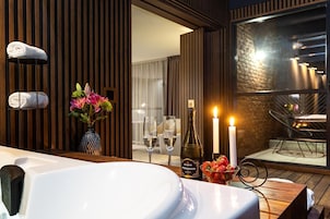 A modern bathroom with a large bathtub, towels, a vase with flowers, two glasses of wine, a bottle of Prosecco, and two lit candles.