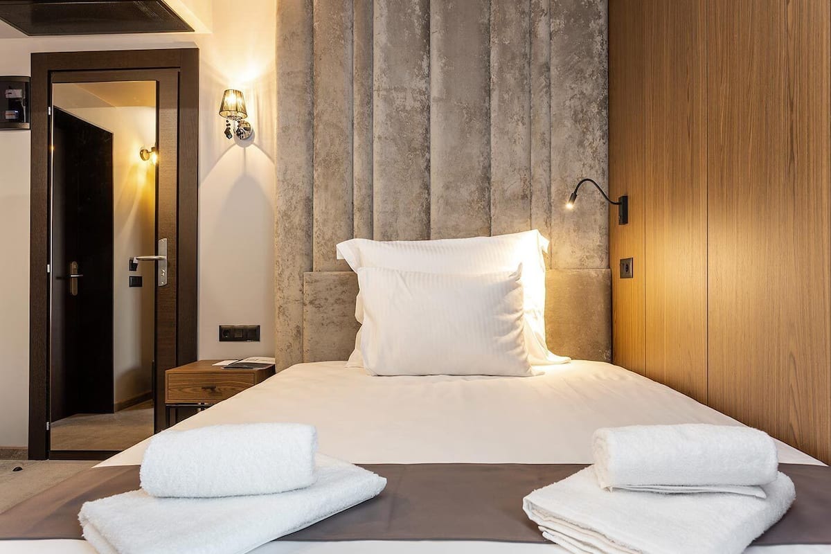 A neatly made bed with white linens and two folded towels, a wooden nightstand with a lamp, and a textured wall behind the bed.