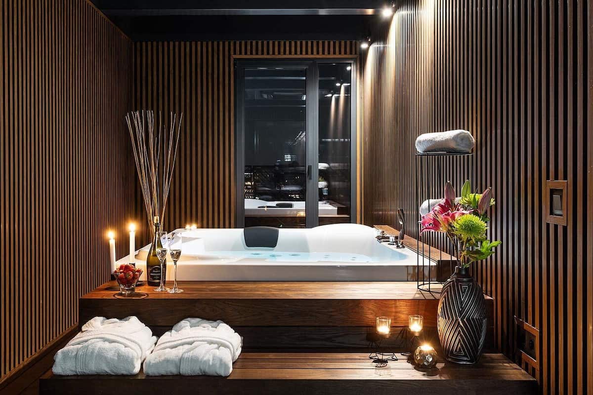 A modern bathroom with a freestanding bathtub, wooden paneling, candles, and a vase with flowers.
