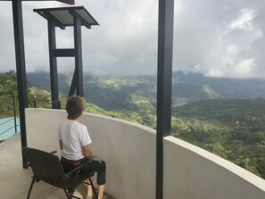 En person sidder i en stol på en balkon med udsigt over et bjergrigt landskab.