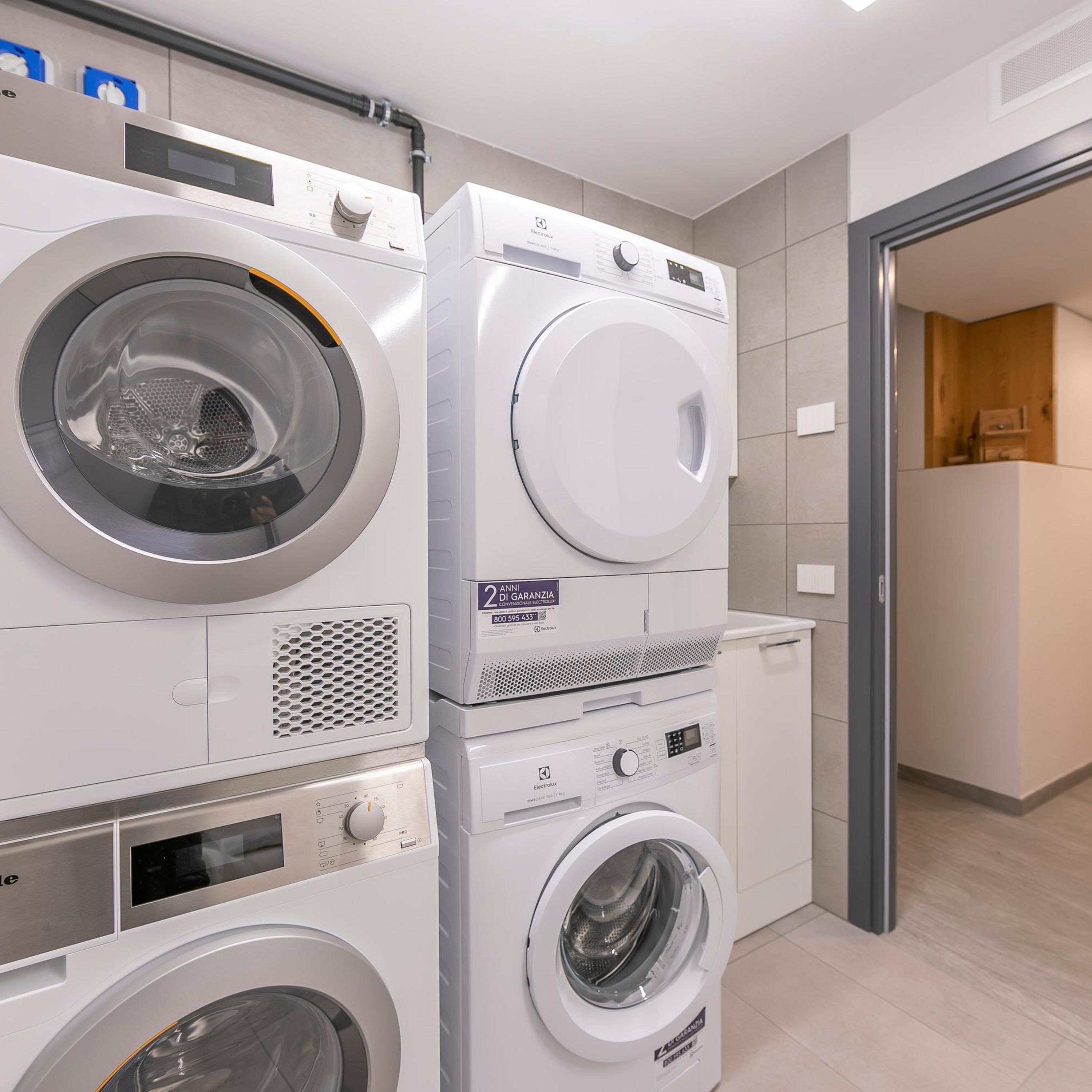 A laundry area with stacked washing machines and dryers, a tiled floor, and a wooden cabinet in the background.
