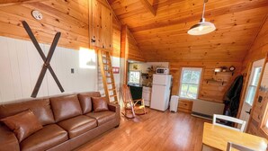 A cozy cabin interior with a wooden interior, a brown leather sofa, a dining table with chairs, and a kitchen area with a refrigerator and stove. - Lakefront Cabin on Rangeley Lake: Sauna & views (Rangeley)
