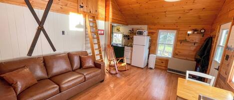 A cozy cabin interior with a wooden interior, a brown leather sofa, a dining table with chairs, and a kitchen area with a refrigerator and stove.
