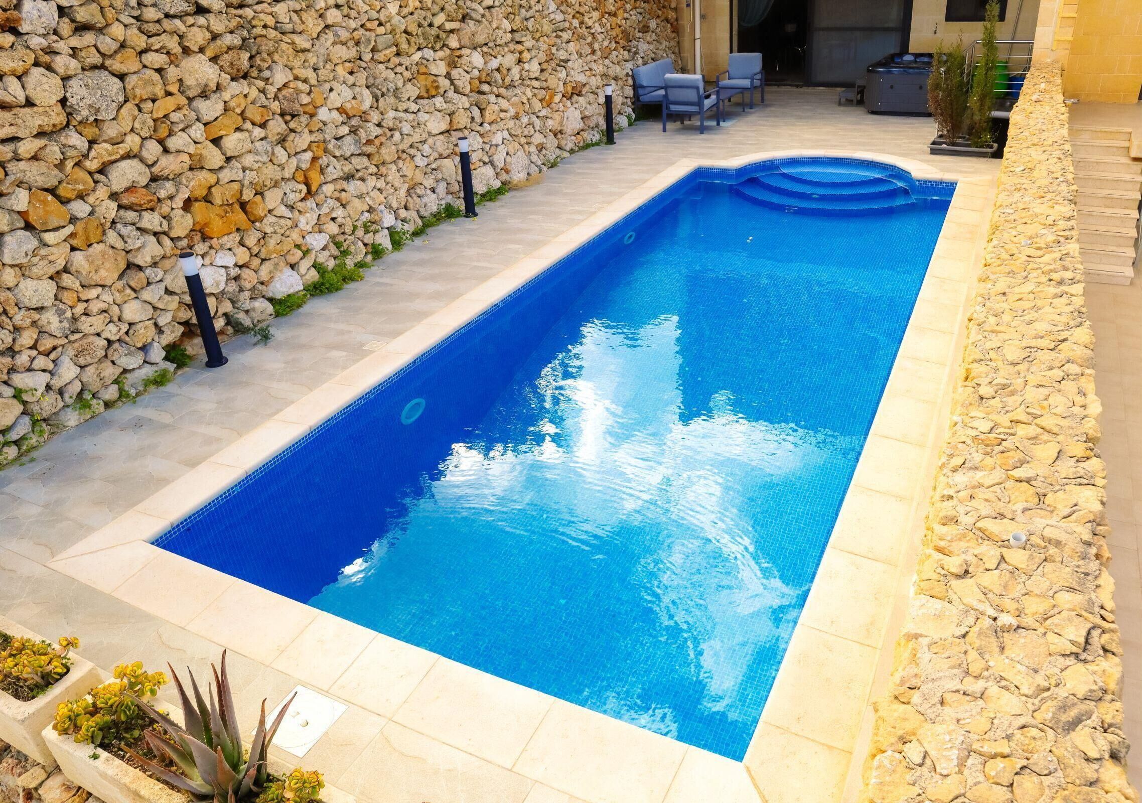 A rectangular swimming pool with a stone wall on one side and a tiled patio area with outdoor furniture.