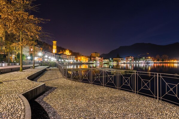 A nighttime riverside walkway with a metal railing, pebbled path, and illuminated buildings in the background.
