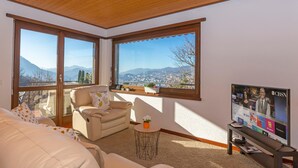 A living room with a large window, a sofa, a chair, and a television displaying a news channel. - Chiara s Home in Pregassona Lugano (Lugano)