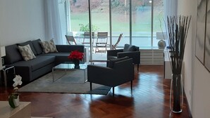 Ein modernes Wohnzimmer mit einer schwarzen Couch, einem Couchtisch und Blick auf eine Grünfläche durch große Fenster.