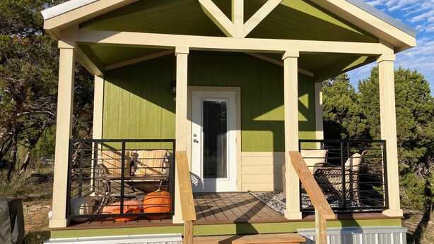 A small, single-story building with a green exterior and a white door. It features a porch with a wooden railing and a small staircase leading up to it. - Cozy Tiny Home in the Hills (Fischer)