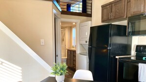 A modern kitchen with a black refrigerator, stove, and microwave. There is a dining table with two chairs and a potted plant. The kitchen has wooden cabinets and a window above the sink. - Cozy Tiny Home in the Hills (Fischer)
