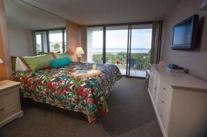 Relax in this serene hotel room featuring a large bed with a floral-patterned bedspread, a flat-screen TV, and a balcony with ocean views.