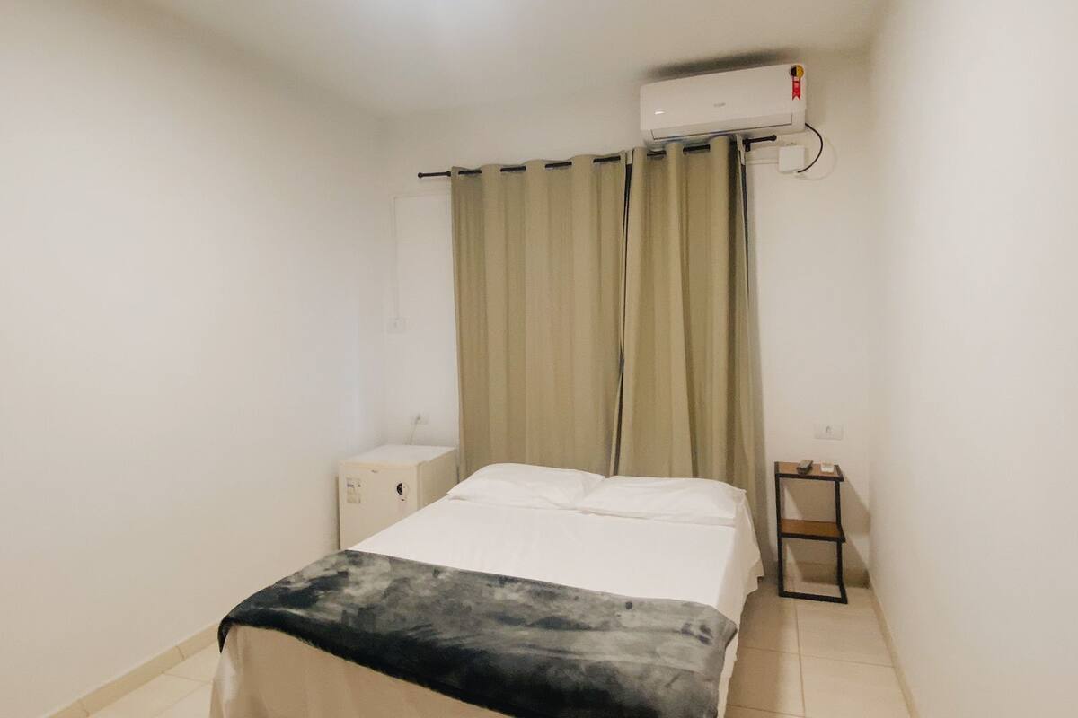 Une petite pièce propre, comprenant un lit, une table de chevet, une commode, un appareil de climatisation et un rideau.