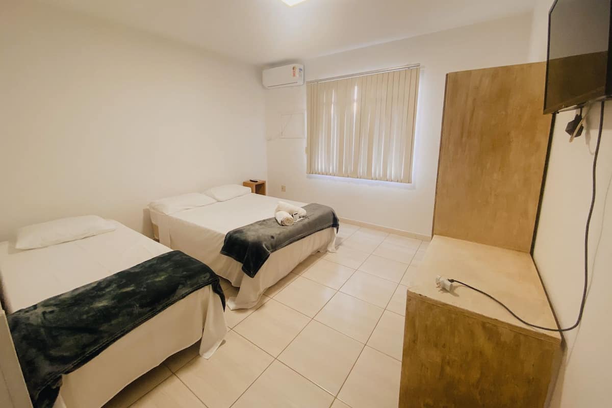 Une chambre d’hôtel avec deux lits simples, une petite table de chevet, une fenêtre avec des stores, un appareil de climatisation et un téléviseur posé sur un meuble en bois.