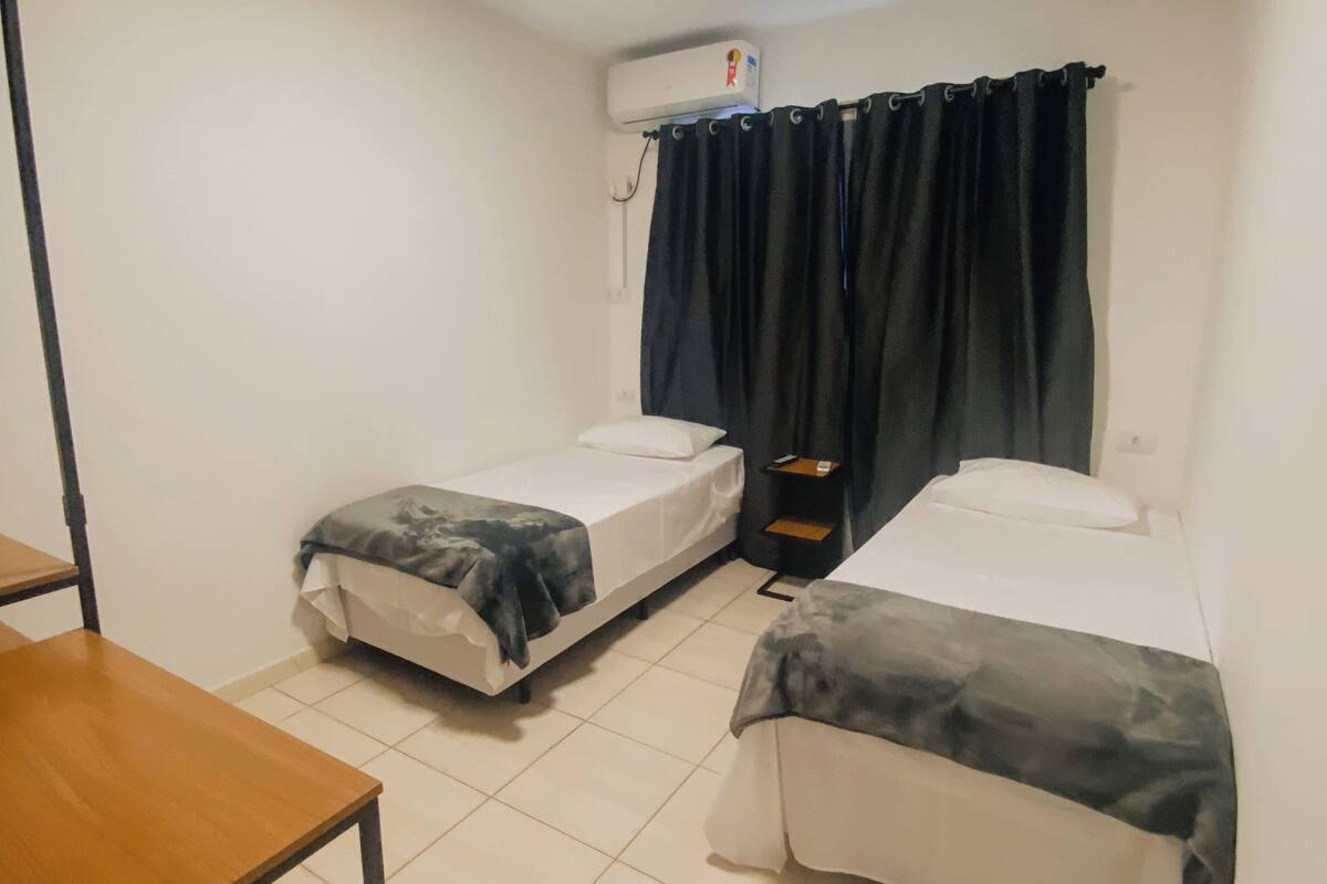 Une petite chambre propre, avec deux lits simples, une petite table et une fenêtre avec des rideaux foncés.