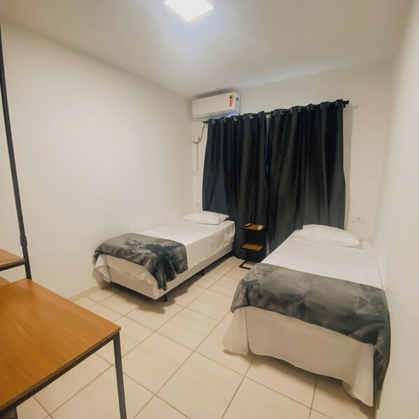 Une petite chambre propre, avec deux lits simples, une petite table et une fenêtre avec des rideaux foncés.