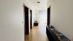Un long couloir étroit, doté d’un plancher en bois et de murs blancs. Des portes se trouvent de part et d’autre, un banc noir est placé au centre, et une longue table avec des magazines se trouve au bout.