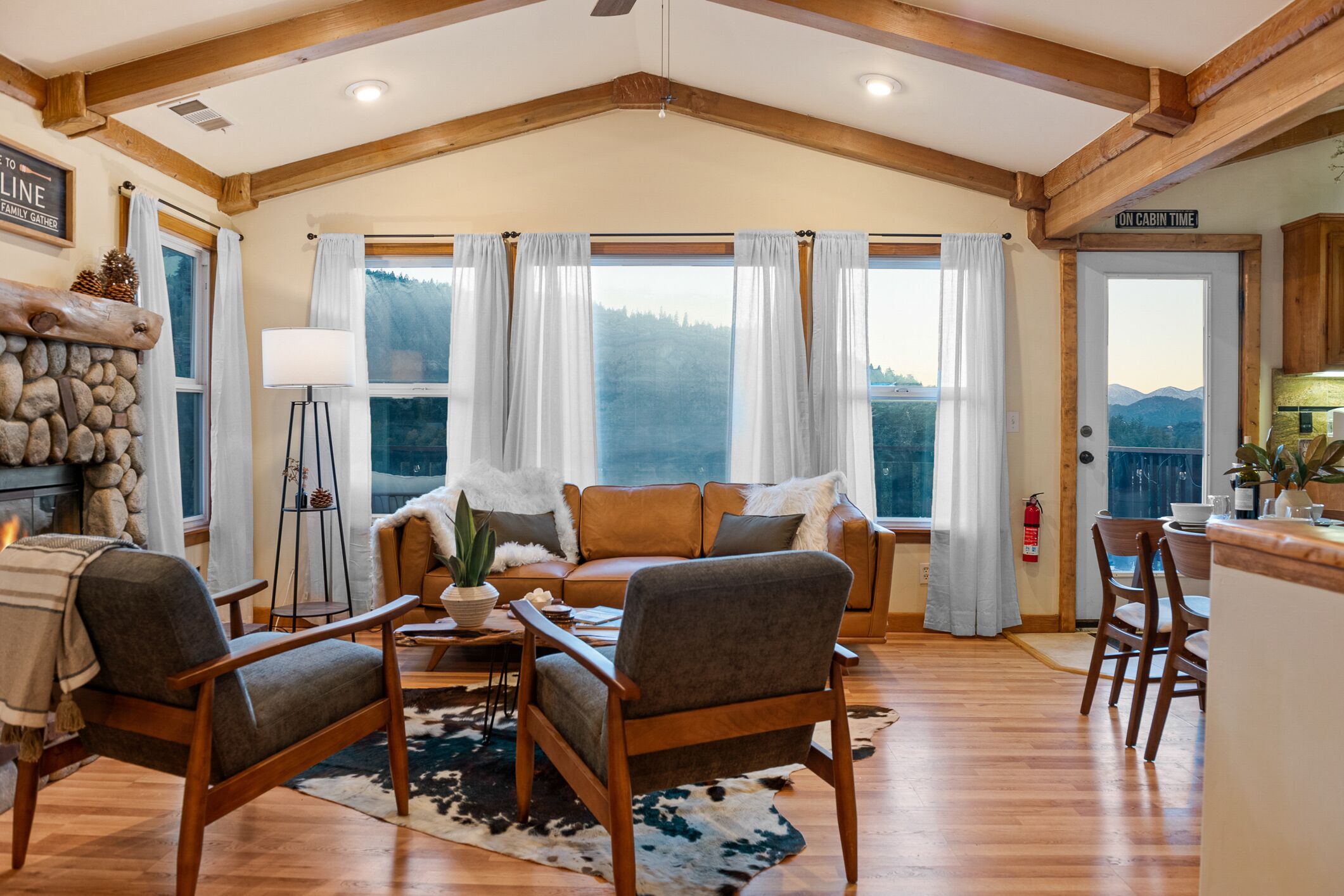 A cozy cabin interior with a fireplace, a sofa set, a dining area, and large windows offering a view of the mountains.
