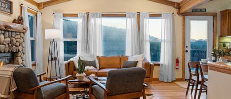 A cozy cabin interior with a fireplace, a sofa set, a dining area, and large windows offering a view of the mountains.
