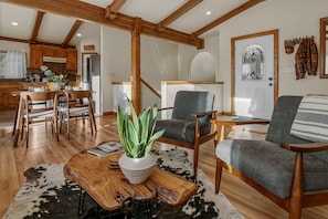 A spacious living room with a wooden coffee table, two grey armchairs, and a potted plant. A dining area with wooden chairs and a kitchen with wooden cabinets is visible in the background.