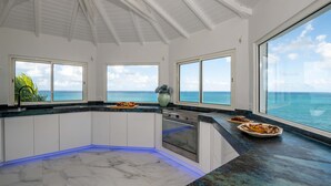 Una cocina moderna con armarios blancos, piso de mármol y grandes ventanales con vistas al océano.