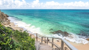 Una escalera de madera que conduce a una playa con aguas turquesas cristalinas y un litoral rocoso.