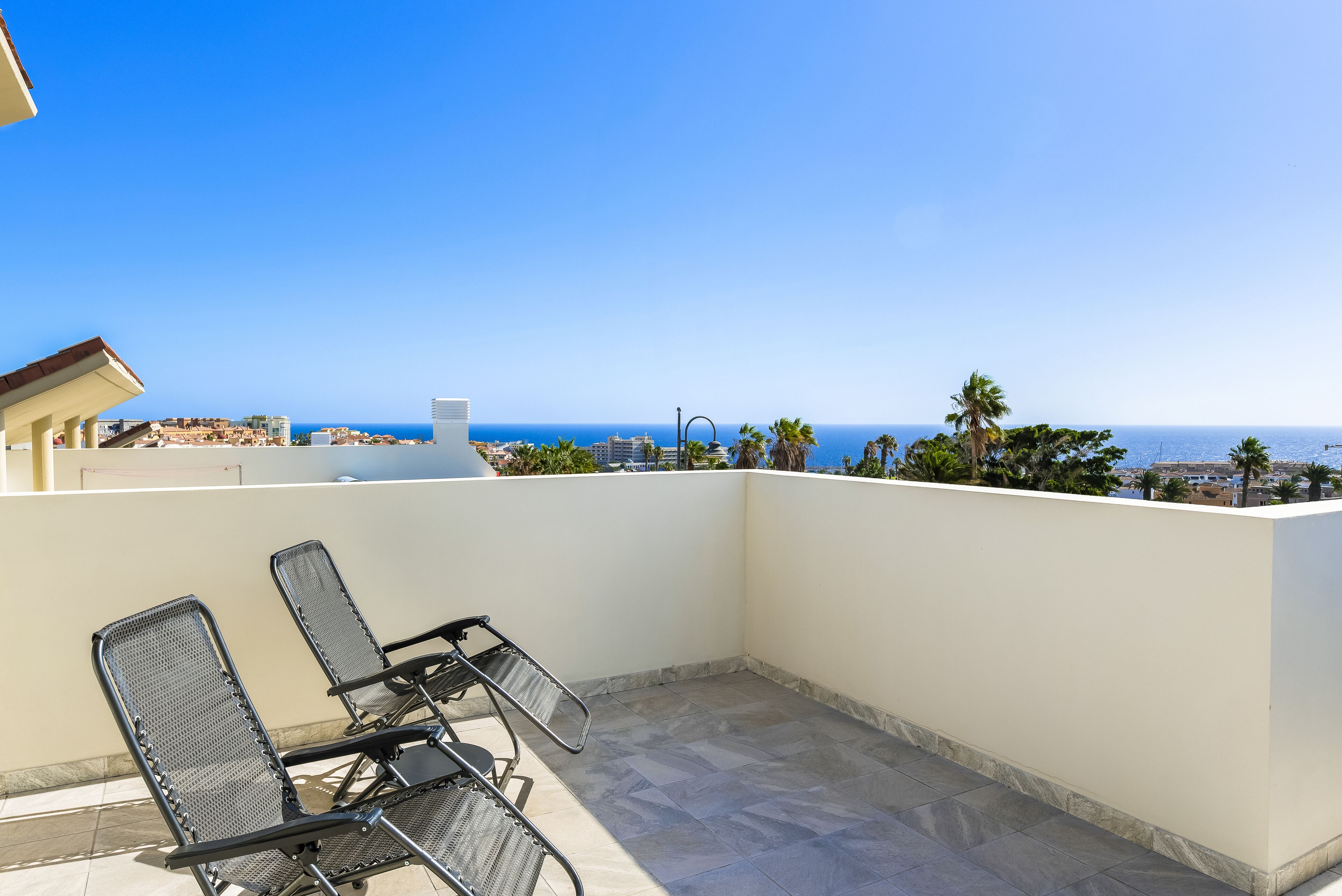 Eine Dachterrasse mit zwei Metallstühlen und Blick auf das Meer sowie Gebäude in der Ferne.