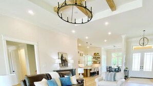 A spacious living room with a brown leather sofa, white armchairs, and a coffee table. A chandelier hangs from the ceiling, and a dining area is visible in the background. - Luxury Farmhouse with Pool - Perfect for Groups (Conroe)