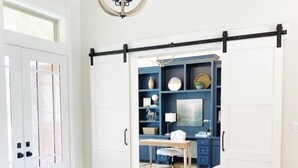 A room with a white door, a sliding barn door, a chandelier, a blue wall with shelves, and a white chair. - Luxury Farmhouse with Pool - Perfect for Groups (Conroe)