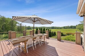 A wooden deck with a table and chairs, an umbrella, and a view of a green field. - Stunning Historic Maine Stone Castle (Bath)