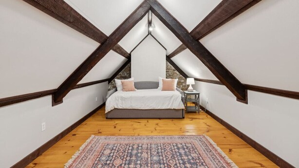 A bedroom with a sloped ceiling, wooden beams, a bed with pillows, a nightstand, and a rug. - Stunning Historic Maine Stone Castle (Bath)