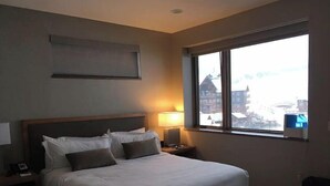 Ein Hotelzimmer mit einem großen Bett, einem Nachttisch mit Lampe, einem Fenster mit Blick auf Gebäude und einer Wand-Klimaanlage.