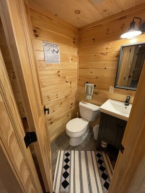 A wooden-paneled bathroom with a toilet, sink, and mirror. - Adirondack Lake Cabins (Indian Lake)