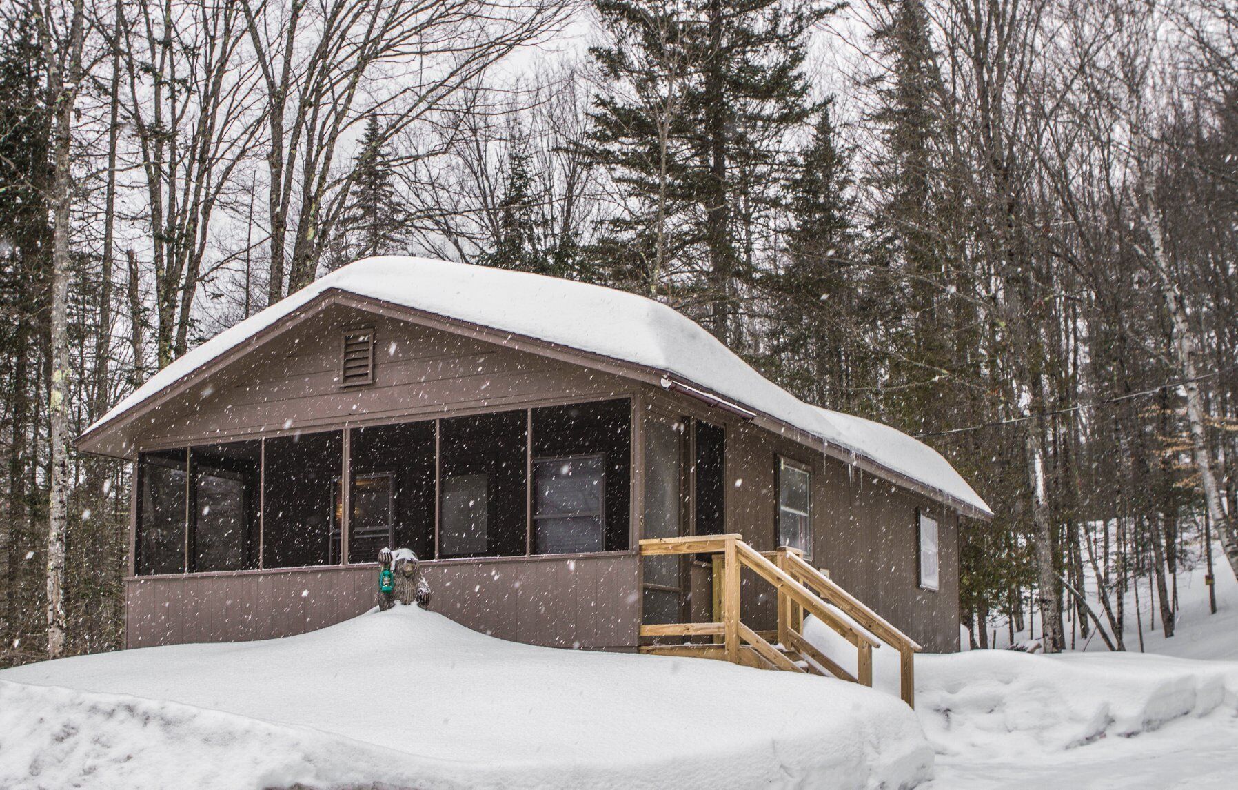 A cabin with a wooden staircase leading to a porch, surrounded by snow-covered ground and trees.
