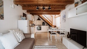 A cozy living room with a sofa, TV, and a dining area with chairs and a table. A kitchen with appliances and a staircase is visible in the background. - Mezzanine apartment in an alpine hamlet (Roure)
