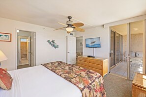 A hotel room with a bed, a ceiling fan, a television, a wooden dresser, and a sliding glass door leading to a balcony. - Ocean View Condo - 1 bedroom Condo in West Maui-  Free WIFI (Lahaina)
