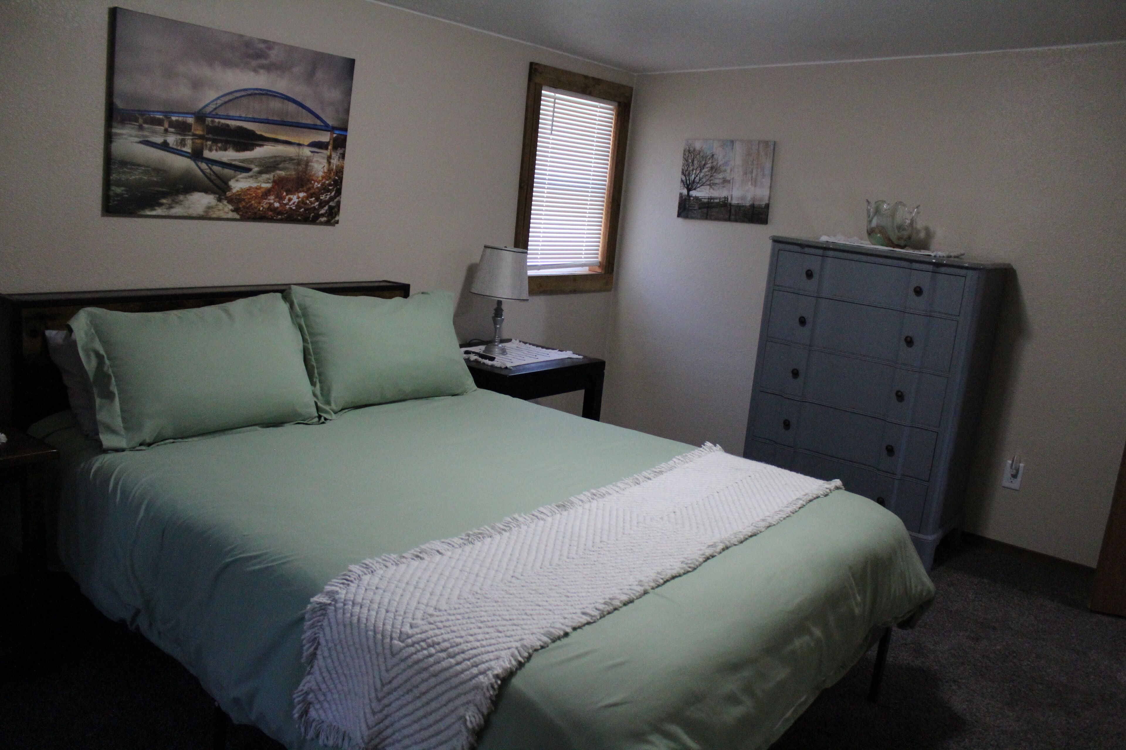A bedroom with a bed, a nightstand, a dresser, a window with blinds, and two wall art pieces.
