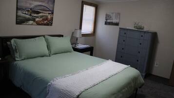 A bedroom with a bed, a nightstand, a dresser, a window with blinds, and two wall art pieces.