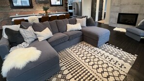 A modern living room with a grey sectional sofa, a patterned rug, and a fireplace. - Premier penthouse  w Private Rooftop near Memorial Stadium & Pinnacle Arena!   (Lincoln)