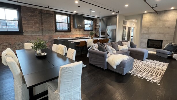 A modern living room with a dark wood floor, a grey sofa, a dining table with chairs, and a kitchen in the background. - Premier penthouse  w Private Rooftop near Memorial Stadium & Pinnacle Arena!   (Lincoln)